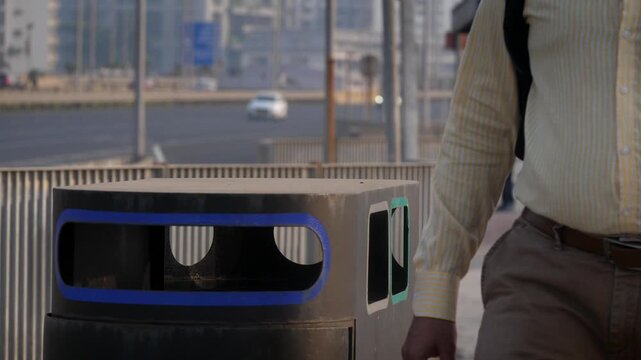 An outdoor daylight shot of a man wearing formal attire and a backpack throws an empty plastic water bottle into a roadside dust bin or trash can with a busy urban street and landscape in the back. 