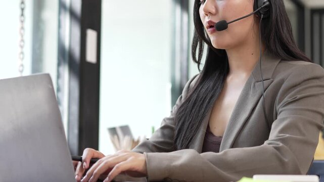 Professional customer service woman wearing a headset typing on a laptop while talking to a client in a modern office call center environment.