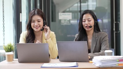 Two smiling Asian female customer service representatives wearing headsets and working on laptops in a modern call center office.