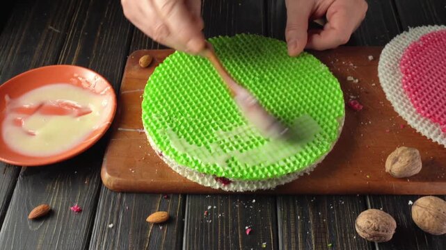A person spreads a condensed milk on a green layered sweet on a wooden board. Nearby, there is a red layer, a bowl with syrup, and some nuts.