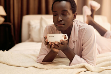 Woman in pajamas relaxing with morning coffee mug on bed