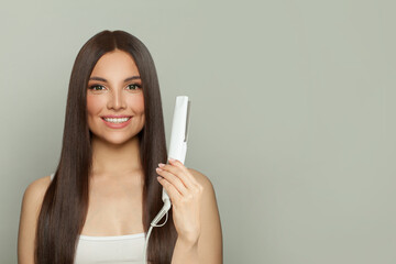 Attractive woman brunette with long straight smooth hairstyle using hair straightener iron on light grey background