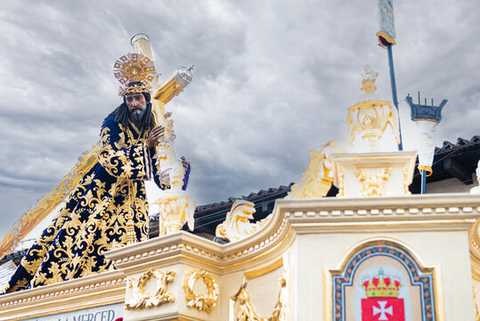 Ornate statue of Jesus with golden crown and embroidered robe, carrying the cross on a float during a Semana Santa procession under cloudy skies.