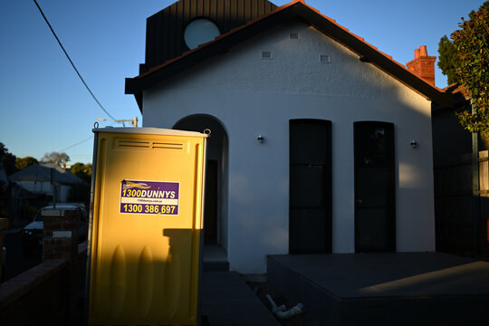 Sunlit yellow portable toilet, from 1300Dunnys, outside a home under renovation late in the day