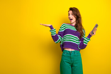 Young woman in striped sweater with phone on bright yellow background smiling and surprised