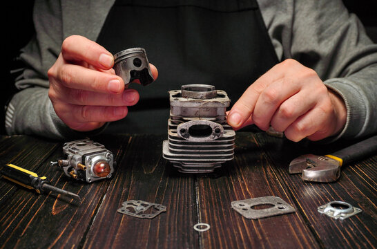 A person holds a small engine part while assembling a larger cylinder on a wooden surface. Tools and other engine components are nearby, showing a focus on repair work