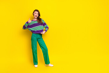 Young female model in striped sweater and green pants smiles against a bright yellow background