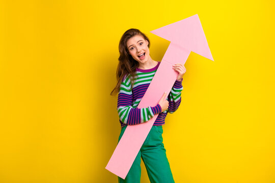 Young woman smiling holds a pink upward arrow against a bright yellow background for fashion lifestyle and promotional mood - Powered by Adobe