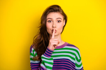 Young woman with finger to lips in striped sweater posing against bright yellow background for...