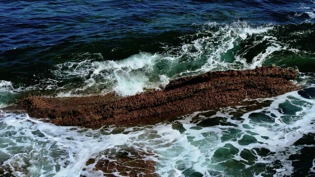 Waves pounding against rugged reef in open ocean. Sea crashing over rough edged coastal rock. Ocean surging along barnacle crusted stone. Surf foaming around jagged seaside formation