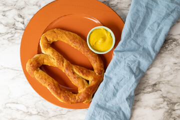 Large, soft pretzel on orange plate, mustard dipping sauce, and blue napkin on marbled surface.