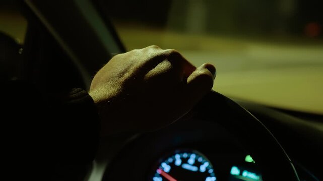 Hand resting on steering wheel while unrecognizable man driving on highway at night. Male cruising road, gripping wheel under street lighting. Nighttime driving along expressway, steering vehicle