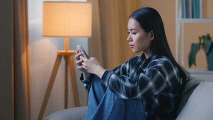 Side view sad Asian woman sitting on sofa at home using smartphone unhappy Korean Chinese female girl scrolling browsing mobile phone app watching social media typing indoors leisure digital cellphone