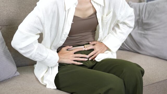 Young Asian woman lying on a sofa holding her stomach in pain, expressing menstrual cramps or abdominal discomfort at home, with a calendar and water glass symbolizing period health.