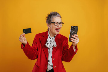 Happy senior woman in stylish red blazer holding smartphone and credit card, celebrating online shopping success against vibrant yellow background