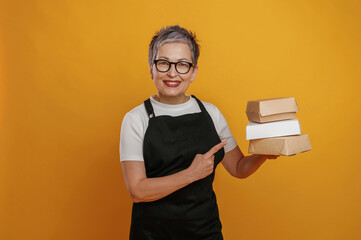 Smiling woman with short gray hair wearing glasses holds stacked takeout boxes against a vibrant yellow background, showcasing food packaging and delivery options