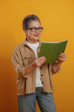 Senior woman with short gray hair is reading a green book while smiling, dressed in casual attire against a vibrant yellow background, showcasing joy of reading
