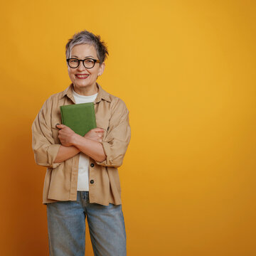 Smiling senior woman with short gray hair wearing glasses holds a green book while standing against a vibrant yellow background, showcasing a joyful reading moment