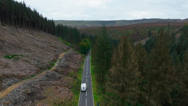 Camper van driving along empty rural road surrounded by trees and hills under cloudy sky. Vehicle moving through scenic valley emphasizing road safety and traffic awareness in remote infrastructure