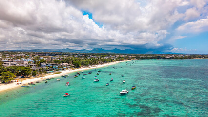 Aerial view of a tropical turquoise lagoon in Mauritius with many boats anchored near a white sand beach and a coastal town under a dramatic cloudy sky