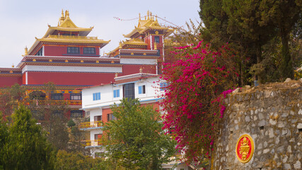 A large traditional monastery with a brick red facade, surrounded by trees and blooming plants, creating a serene and cultural setting.