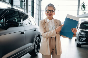 Confident businesswoman in stylish suit is presenting a blue folder while standing beside luxury cars in a modern dealership, showcasing professionalism and success
