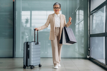 Senior woman in stylish beige suit is walking through modern airport terminal, pulling suitcase and carrying shopping bags, embodying travel and leisure lifestyle