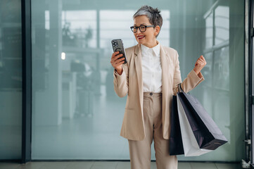 Stylish older woman in beige suit is happily checking her phone while holding shopping bags, showcasing modern fashion and consumer lifestyle in a bright environment