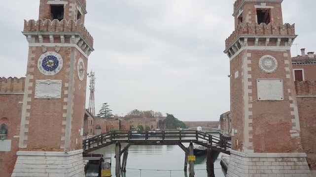 Venetian Arsenal entrance towers with wooden bridge over canal. Historic brick architecture in Venice Italy. Ancient shipyard gateway with clock and sundial under overcast sky outdoors.