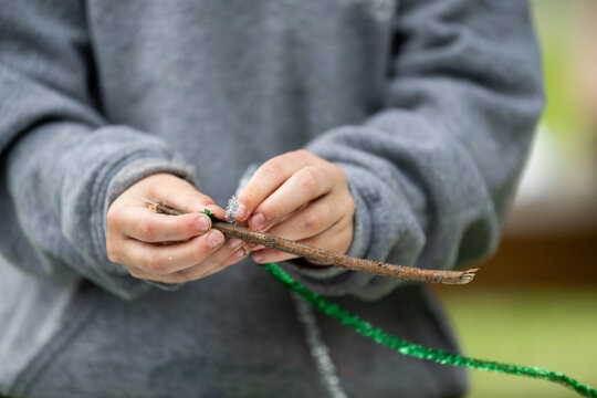 Close-up view captures a child's small hands carefully wrapping a sparkly green pipe cleaner around a wooden stick during an arts and crafts project. 