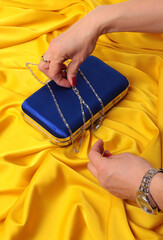 Elegant blue satin clutch bag with silver chain on vibrant yellow silk background. Woman's hands with red nails holding a luxury evening purse. High-contrast fashion lifestyle product photography.