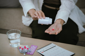 Woman taking medication at home, pouring a capsule into her hand beside a glass of water, blister...