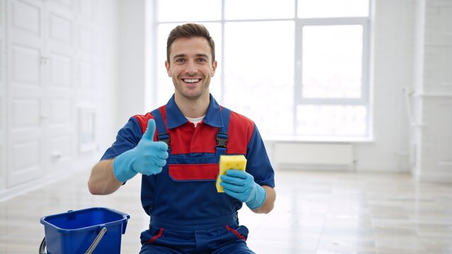 Smiling professional cleaner in uniform showing a thumbs up. Happy young male janitor with a sponge and bucket after cleaning a room. Quality housekeeping service concept with copy space - Powered by Adobe