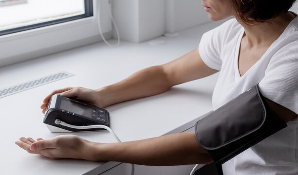 Close-up of a person checking blood pressure at home with an automatic digital monitor on a white table near the window, concept of self-care and hypertension control