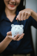 An Asian businesswoman smiles as she drops coins into a piggy bank at her desk, demonstrating smart...