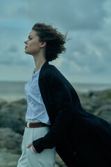 Wind swept profile on a rocky shore, wearing a white blouse, belt and dark coat, embodying calm...