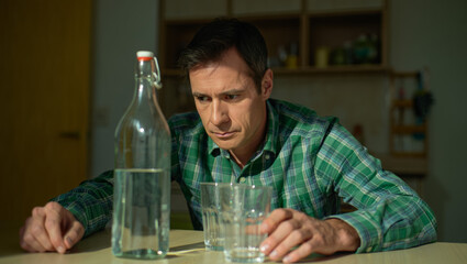 A man in a green checkered shirt sits alone at a table with a glass and a bottle of water, appearing contemplative in a softly lit, quiet home interior.