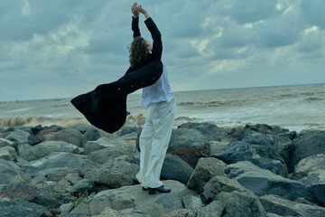 Dancer on rocky shore spins and leaps as wind lifts a flowing coat, white pants and dark jacket against a moody seascape, creating dynamic movement, freedom, and dramatic energy.