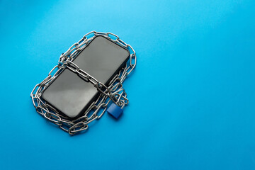 High angle of a smartphone wrapped in metal chains and secured with a blue padlock on blue background. Concept for technology addiction, internet slavery, digital detox, and cyber security.