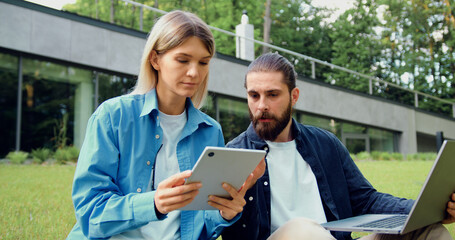 Two colleagues sitting on the grass outdoors, collaborating with a laptop and a tablet. The man explains something to the woman, pointing to the screen on her tablet as they work together.