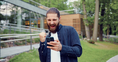 Surprised Caucasian man standing in a city park, using a smartphone and reacting emotionally to unexpected news. Concept of online communication and technology.