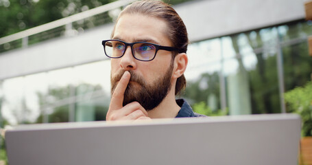 Close-up of a focused bearded businessman in glasses watching a monitor screen, outdoors. Professional concentration and modern technology concept
