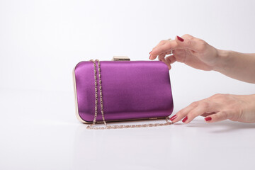 Elegant purple satin clutch bag with gold chain strap. Woman's hands with red nails reaching for a luxury evening purse isolated on white background. Professional fashion accessory photography.