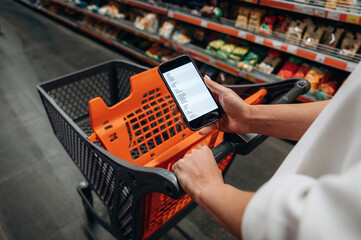 Person holding smartphone while pushing shopping cart in grocery store aisle, surrounded by shelves filled with various food products and items © standret