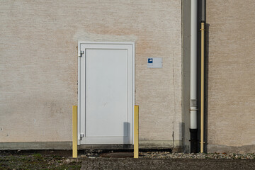 A small white door stands against a plain wall in an urban setting. Yellow poles are positioned in front of the door, and a sign is visible nearby. The scene shows daily city life.