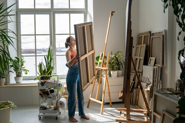Talented woman artist painting at home in spacious loft with houseplants and panoramic windows. Creative female painter replacing artwork canvas on easel, practicing art hobby and dedication to craft.