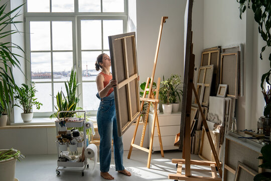 Talented woman artist painting at home in spacious loft with houseplants and panoramic windows. Creative female painter replacing artwork canvas on easel, practicing art hobby and dedication to craft.