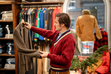 Obraz premium Serious mustachioed man shopping for outerwear in second hand vintage store, deciding on buying chic wool jacket, male in shop holding clothing on hanger and looking at model and quality of fabric.
