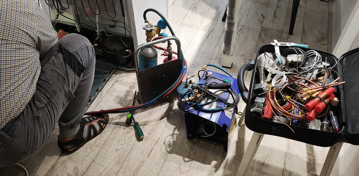 Person repairing refrigerator compressor with gas tanks, refrigeration tools, and copper pipes, professional service technician performing maintenance.