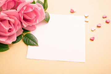 Beautiful pink roses with blank white greeting card and small heart confetti on beige background